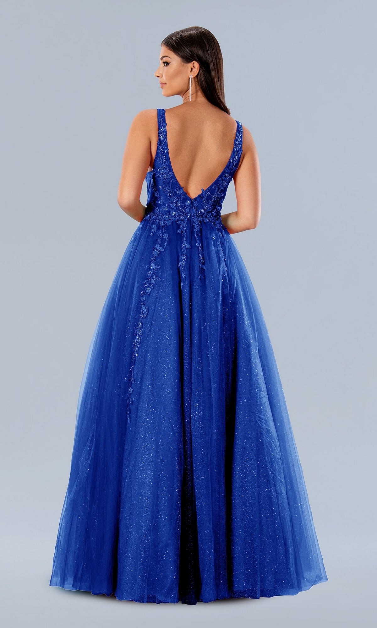 Woman wearing the Stella Couture 24203 Long Glitter Prom Gown, a sleeveless, floor-length royal blue dress with a deep V-back, standing against a plain light gray background.